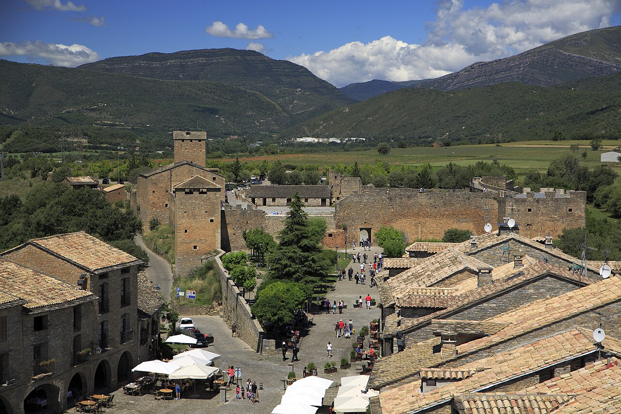 Aínsa vista desde lo alto: la plaza Mayor porticada con terrazas en primer término, el recinto amurallado del castillo al fondo y, detrás, el valle verde y las sierras del Sobrarbe bajo un cielo con nubes.