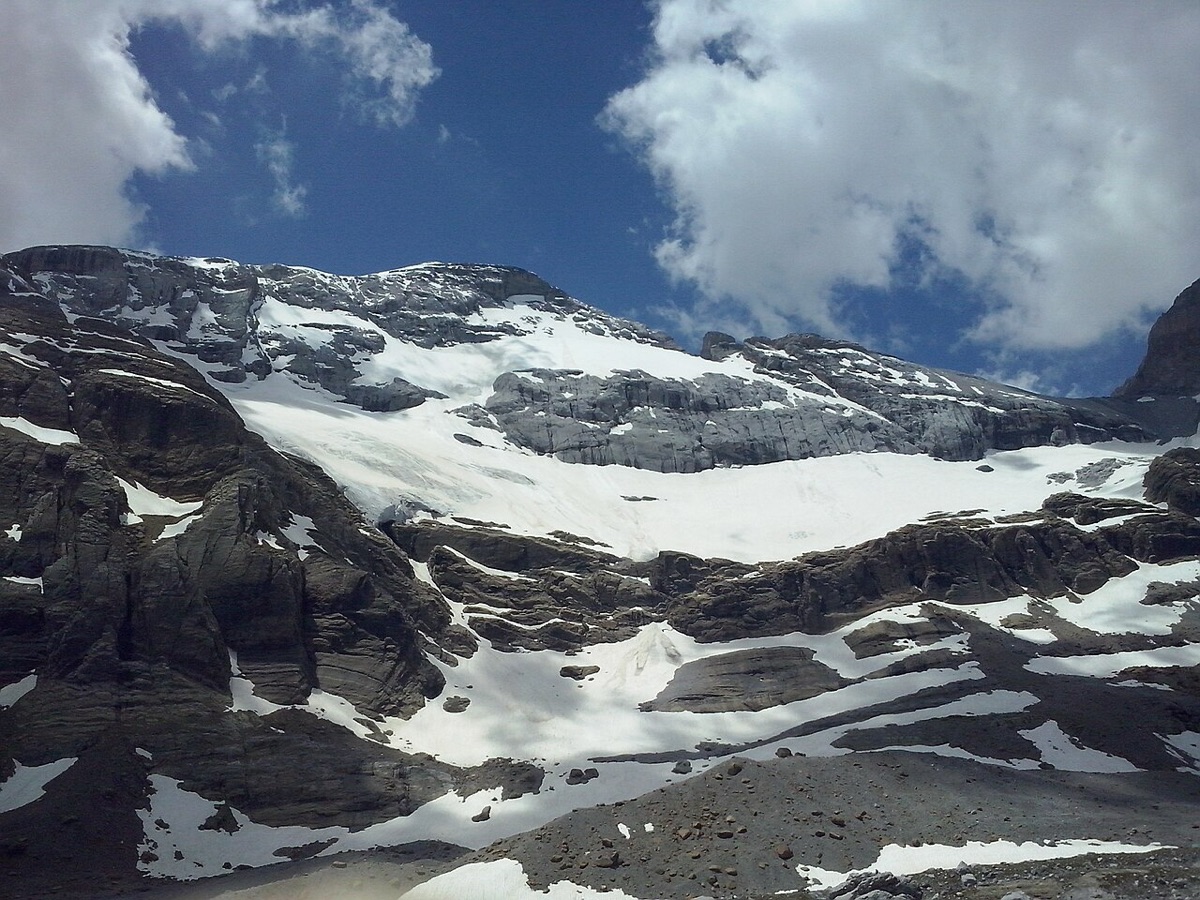 El glaciar del Monte Perdido visto desde el balcón de Pineta: una lengua de hielo y nieve encajada entre bandas horizontales de caliza gris, con neveros sueltos y pedreras en primer término bajo un cielo de nubes de tarde.