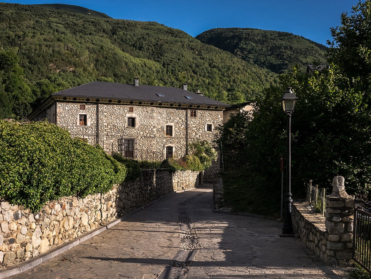 Una calle empedrada y vacía de Anciles subiendo entre muros de piedra, con una casa solariega de sillar y tejado de pizarra al fondo y la ladera boscosa del valle de Benasque detrás.
