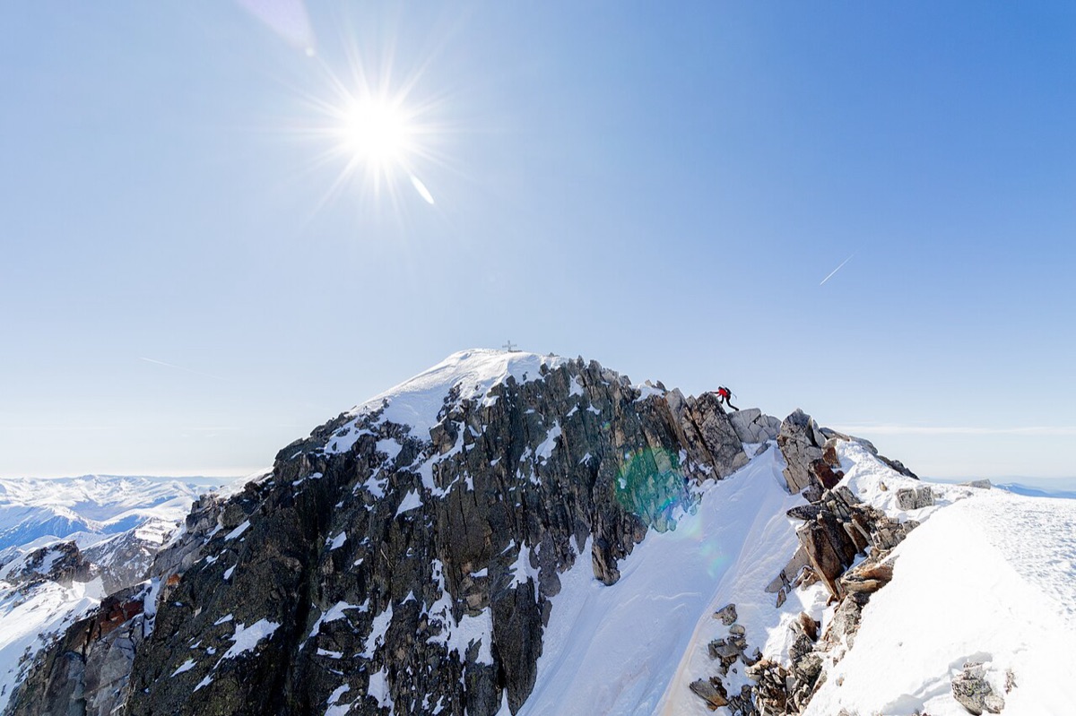 Un montañero con chaqueta roja cruzando el Paso de Mahoma del Aneto: una cresta de bloques de granito con vacío a los dos lados, y al fondo la cúpula nevada de la cima con su cruz metálica recortada contra el cielo.