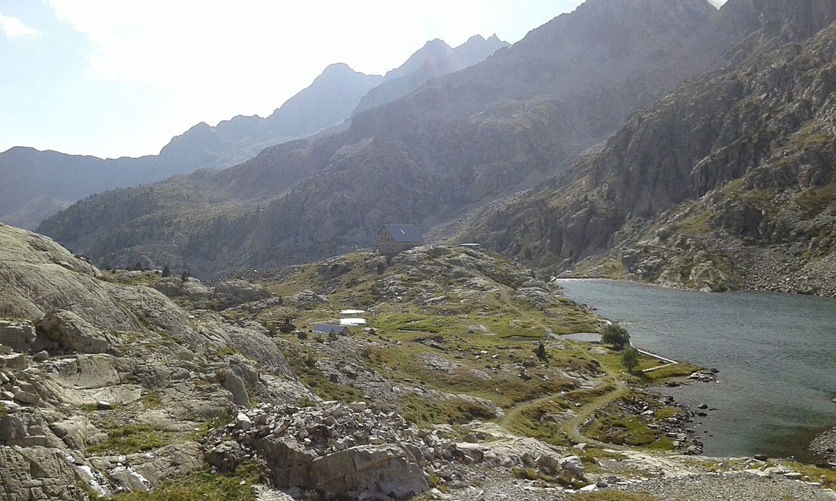 El ibón bajo de Bachimaña con el agua verde azulada ocupando la derecha de la imagen, el refugio de los Ibones de Bachimaña sobre una loma de hierba, y losas de granito pulido en primer término bajo las crestas del valle.