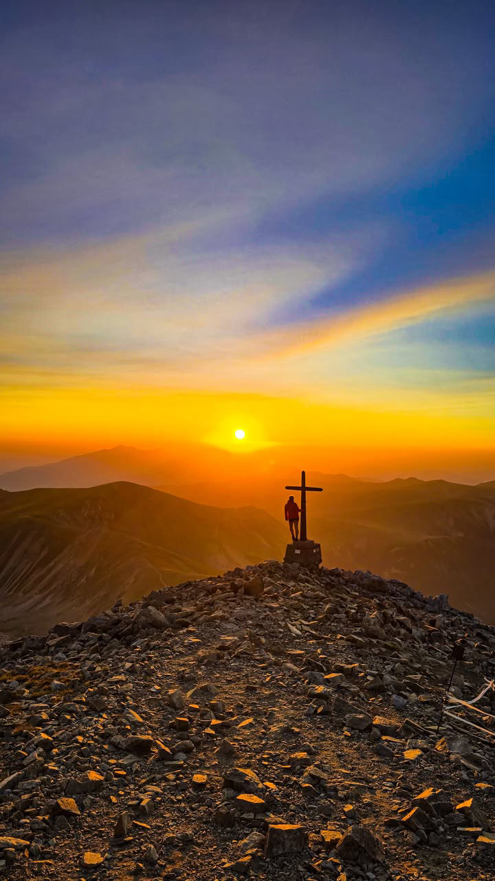 Amanecer desde el Bastiments: el sol asoma naranja sobre un mar de sierras azules y una persona, de espaldas, apoyada en la cruz metálica que hay unos metros bajo la cima, en medio de un pedregal dorado por la primera luz.