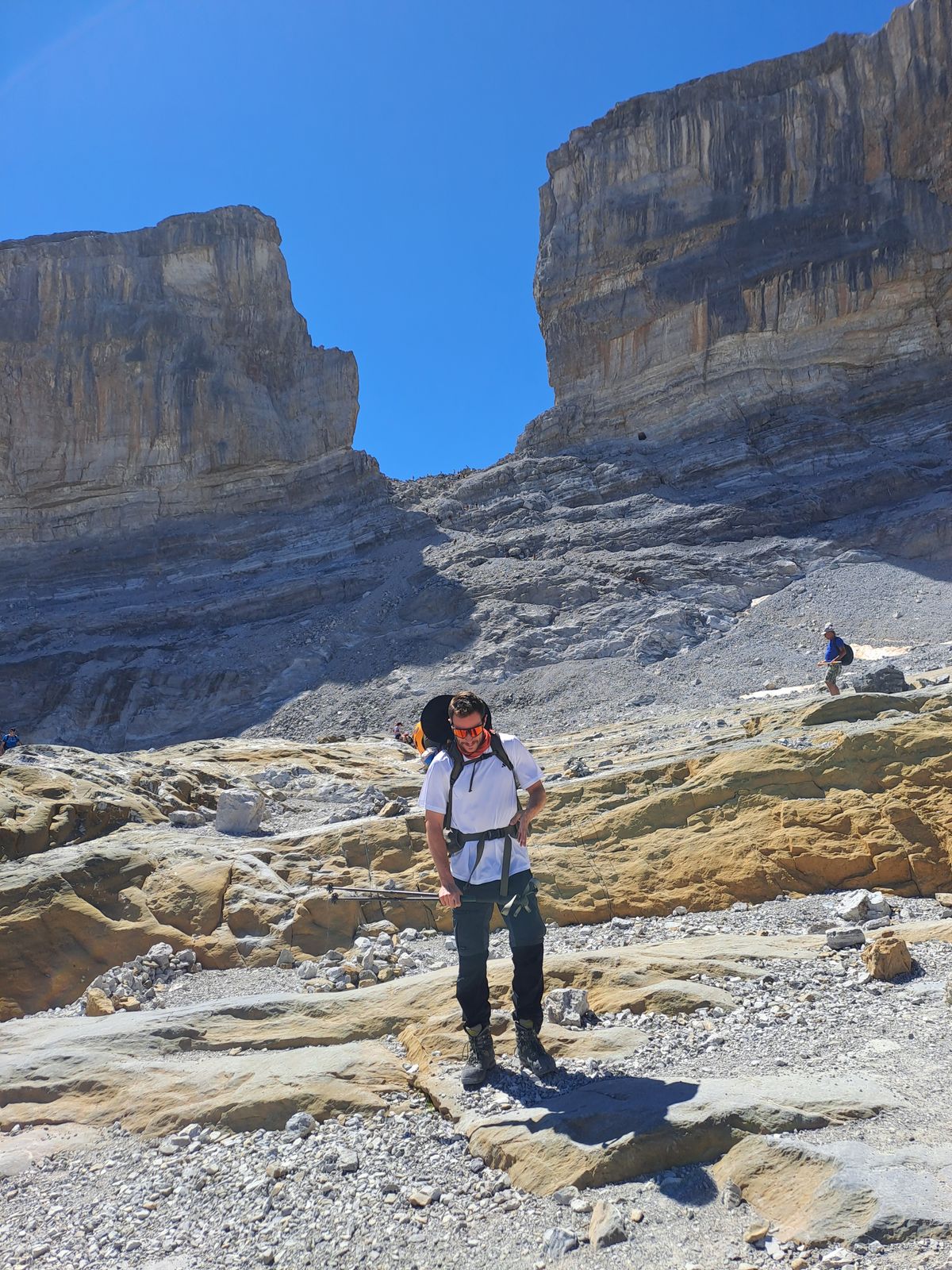 Un montañero con camiseta blanca y mochila se detiene en un pedregal gris bajo la Brecha de Rolando: un hueco vertical de 40 metros de ancho entre dos murallas de roca caliza, con el cielo azul asomando entre ellas.