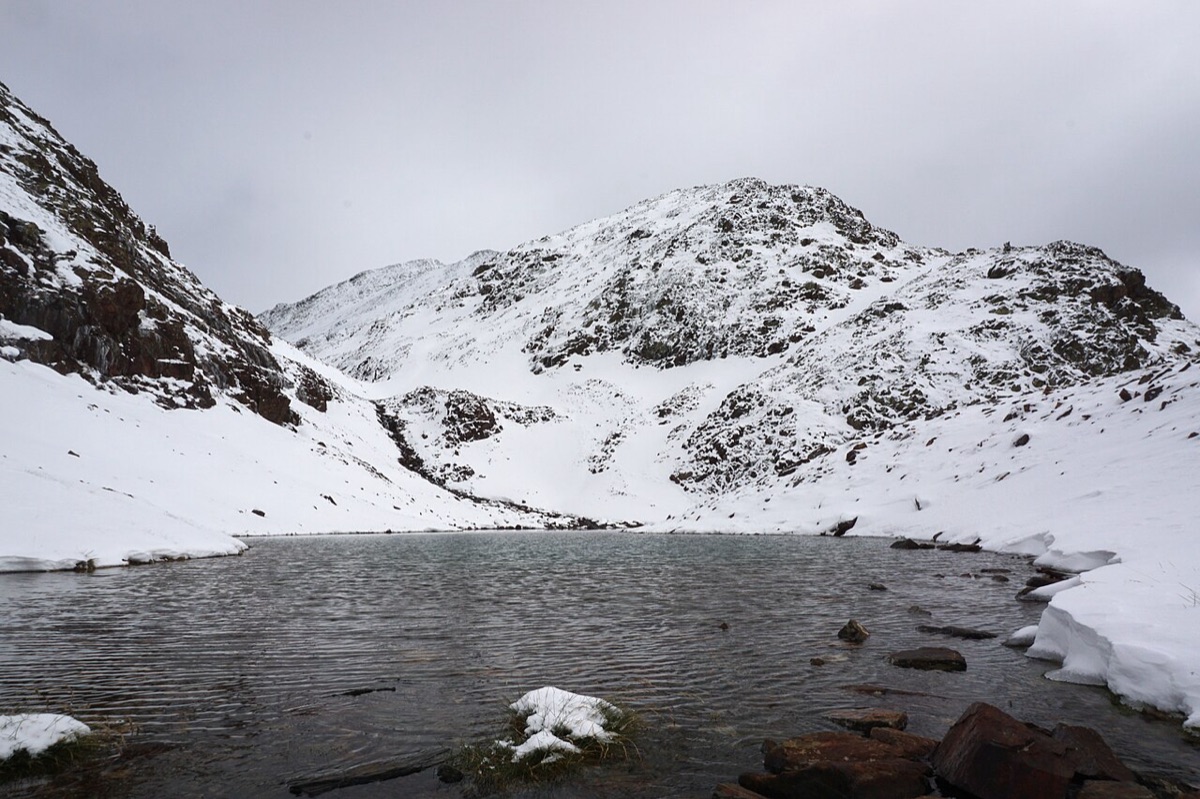 Una de las basses del Estany Negre, en el parque natural del Comapedrosa, con la primera nieve del año: el agua oscura en primer término y las laderas y la cima cubiertas de blanco bajo un cielo encapotado.