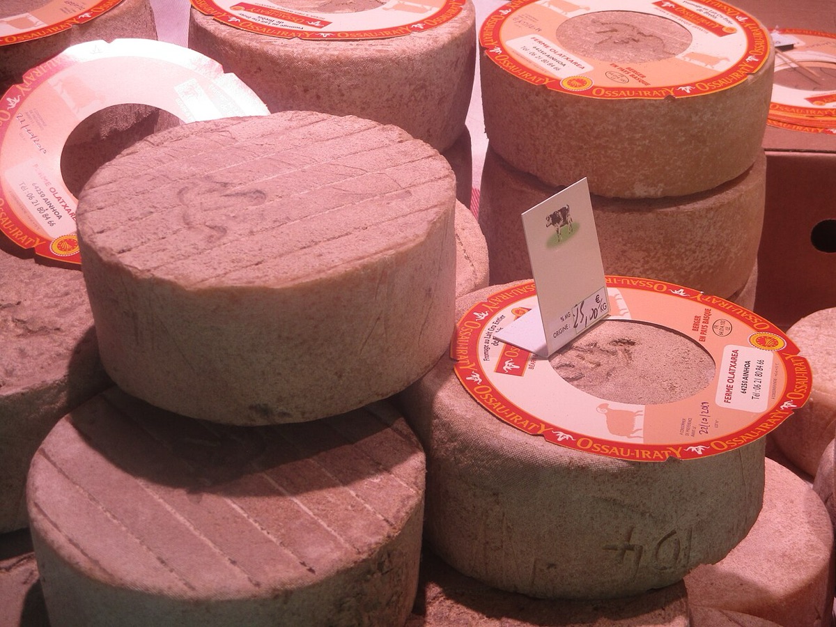 Un montón de ruedas de queso Ossau-Iraty apiladas en un puesto de mercado, con la corteza natural gris y rugosa y la banda roja de la denominación de origen rodeando cada pieza.