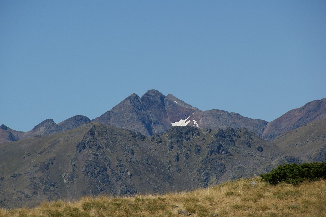 La Pica d'Estats vista de lejos: tres cimas casi iguales recortadas sobre un cielo azul limpio, con un nevero colgado en la cara sur y, delante, cordales de hierba seca y roca oscura.