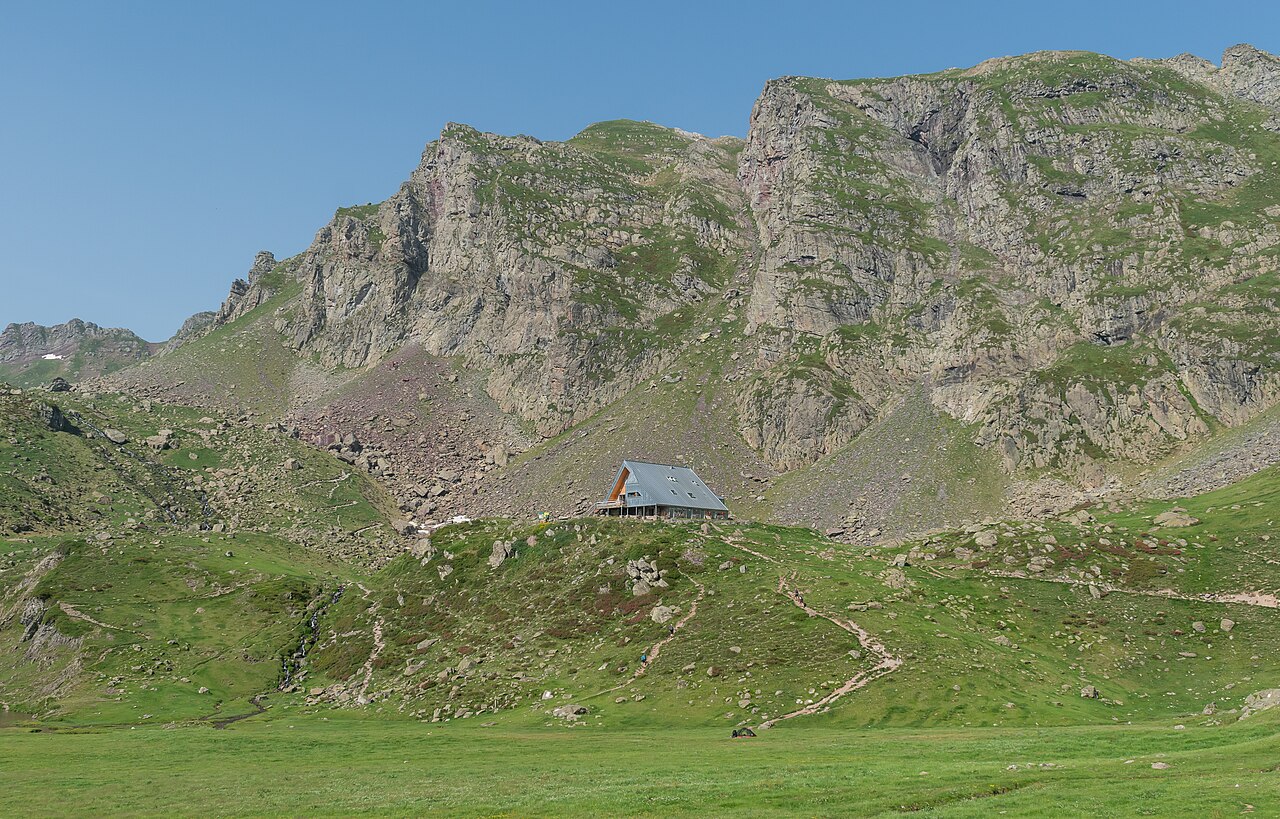 El refugio de Ayous, un edificio moderno de tejado inclinado y fachada de madera, solo en una loma verde bajo una cresta de roca y hierba, con un cielo azul sin nubes.