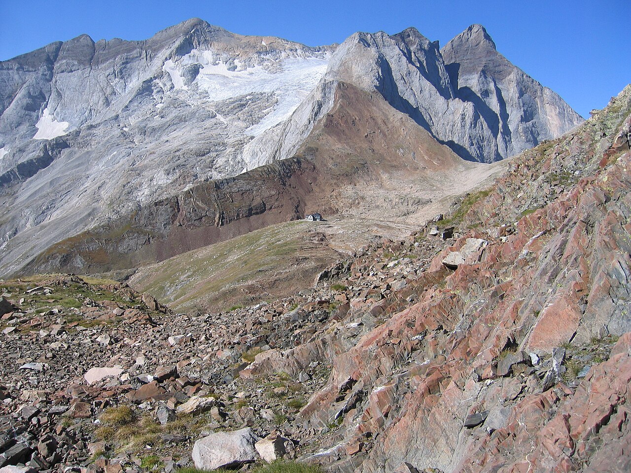 Una ladera de piedra rojiza y grava en primer plano y, al fondo, el refugio de Bayssellance, diminuto sobre una loma, bajo la gran pared gris del Vignemale con su glaciar colgado.