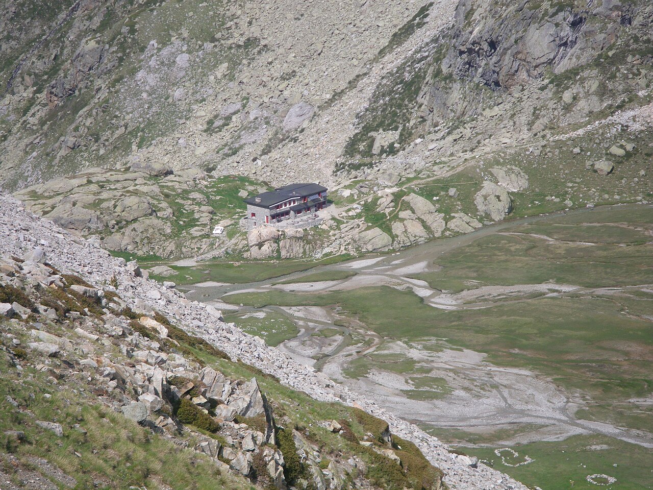 Vista desde arriba del refugio de las Oulettes de Gaube, un edificio de piedra con tejado oscuro sobre un resalte de roca, al borde de una vega verde y pedregosa y bajo laderas de bloques.