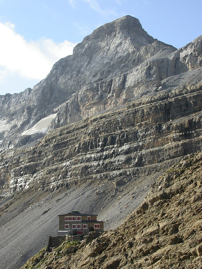 El refugio de los Sarradets, un pequeño edificio de piedra con ventanas rojas, pegado a la ladera de grava bajo un enorme muro de caliza estratificada rematado por una cima de roca.