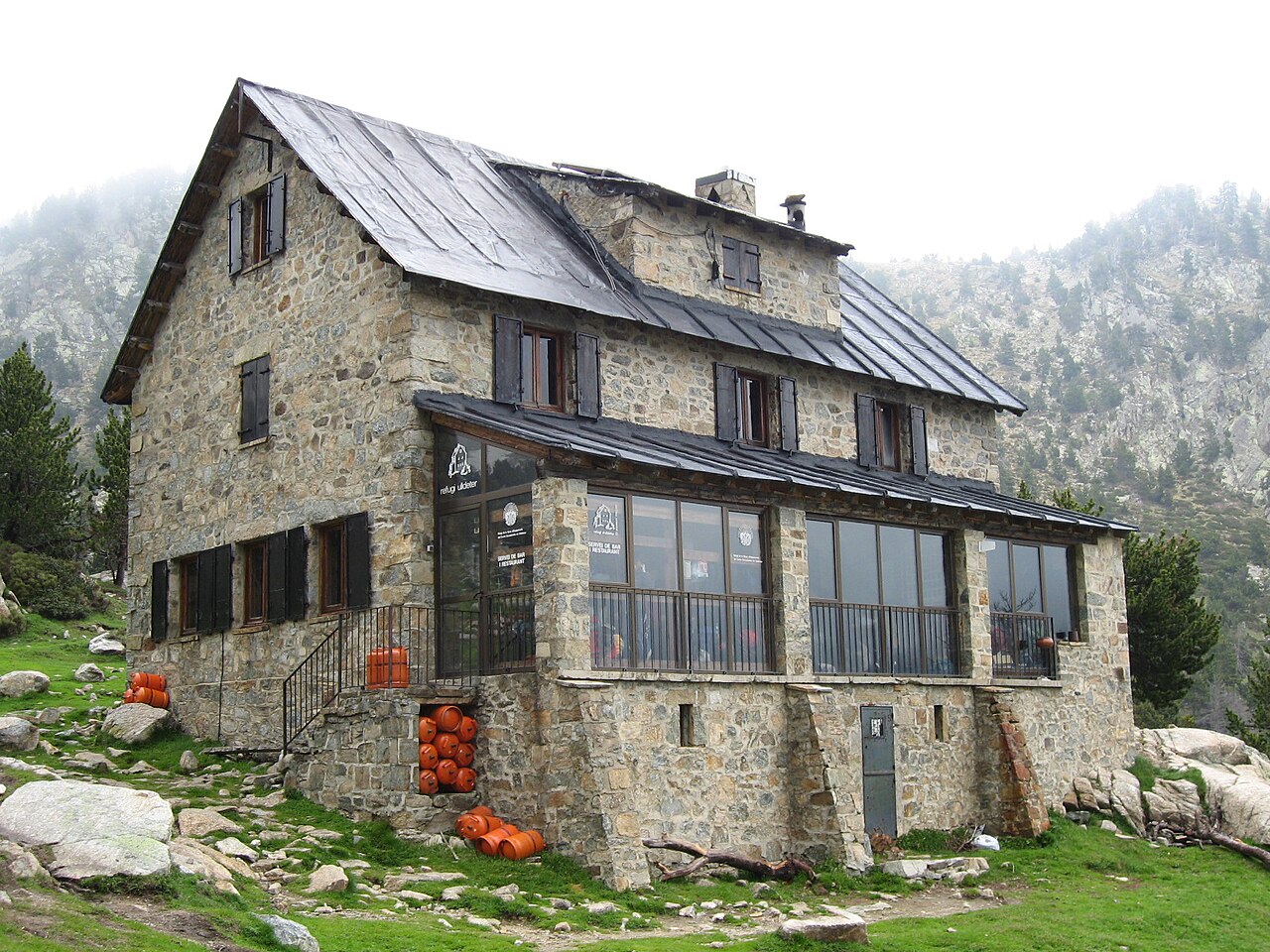 El refugio de Ulldeter, un gran edificio de piedra con tejado oscuro y una galería acristalada con cartel de bar y restaurante, entre pinos y laderas de roca con niebla.