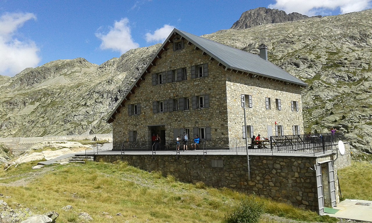 El refugio de los ibones de Bachimaña, un gran edificio de piedra de tres alturas con tejado de chapa y una terraza con barandilla donde hay excursionistas sentados, bajo laderas de granito y un cielo azul con nubes.