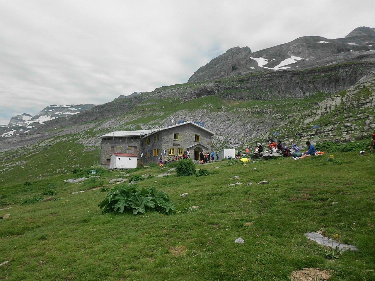 El refugio de Góriz, un edificio de piedra con ventanas amarillas sobre una ladera de hierba, con excursionistas sentados al sol delante y la ladera caliza del macizo de Monte Perdido detrás.