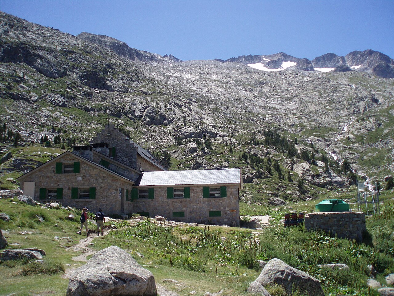 El refugio de la Renclusa, un edificio de piedra con postigos verdes y tejado de pizarra, sobre una ladera de hierba y roca, con dos excursionistas pasando delante y las crestas de la Maladeta con restos de nieve al fondo.